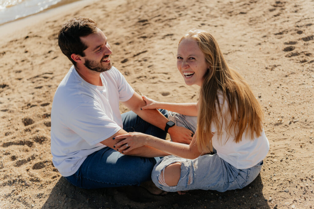 Couple seated in front of the water in the sand and holding arms and laughing